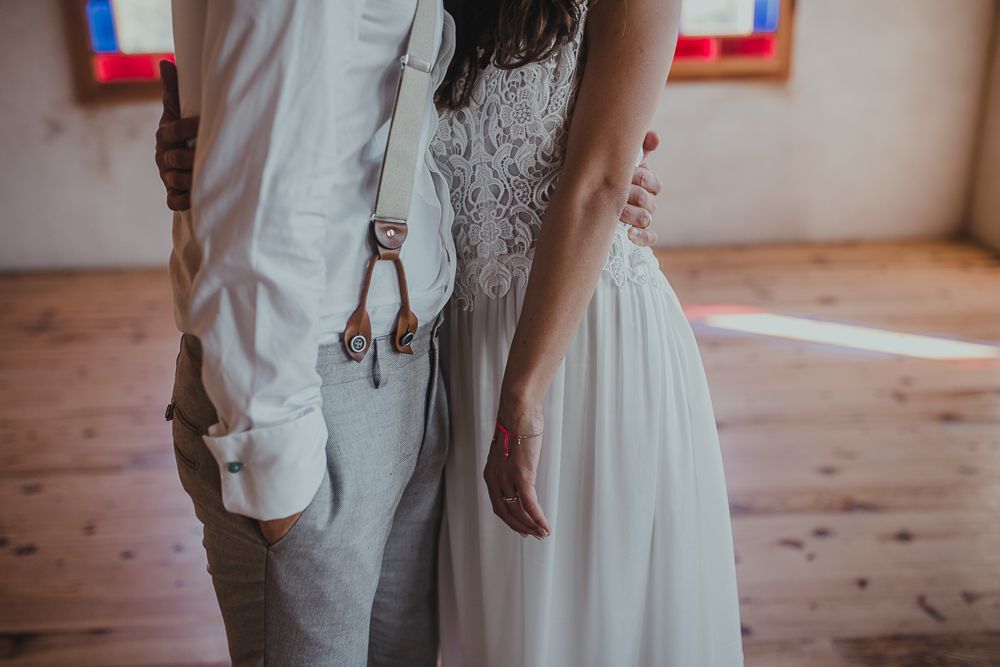 Wedding attire detail showing bride in lace dress and groom in suspenders standing together on wooden floor.