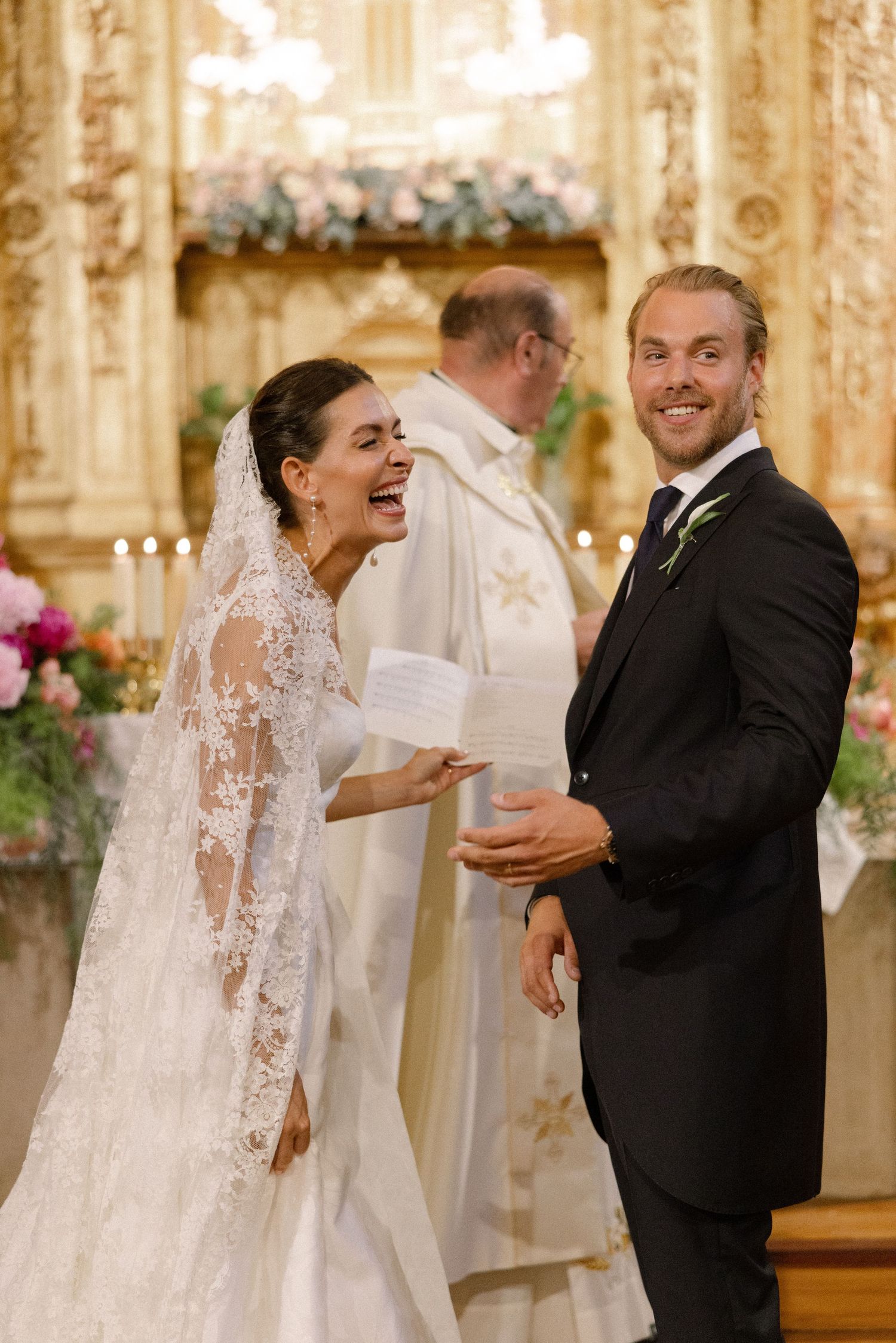 Bride and groom laughing joyfully at altar during elegant church wedding ceremony with ornate golden decor.