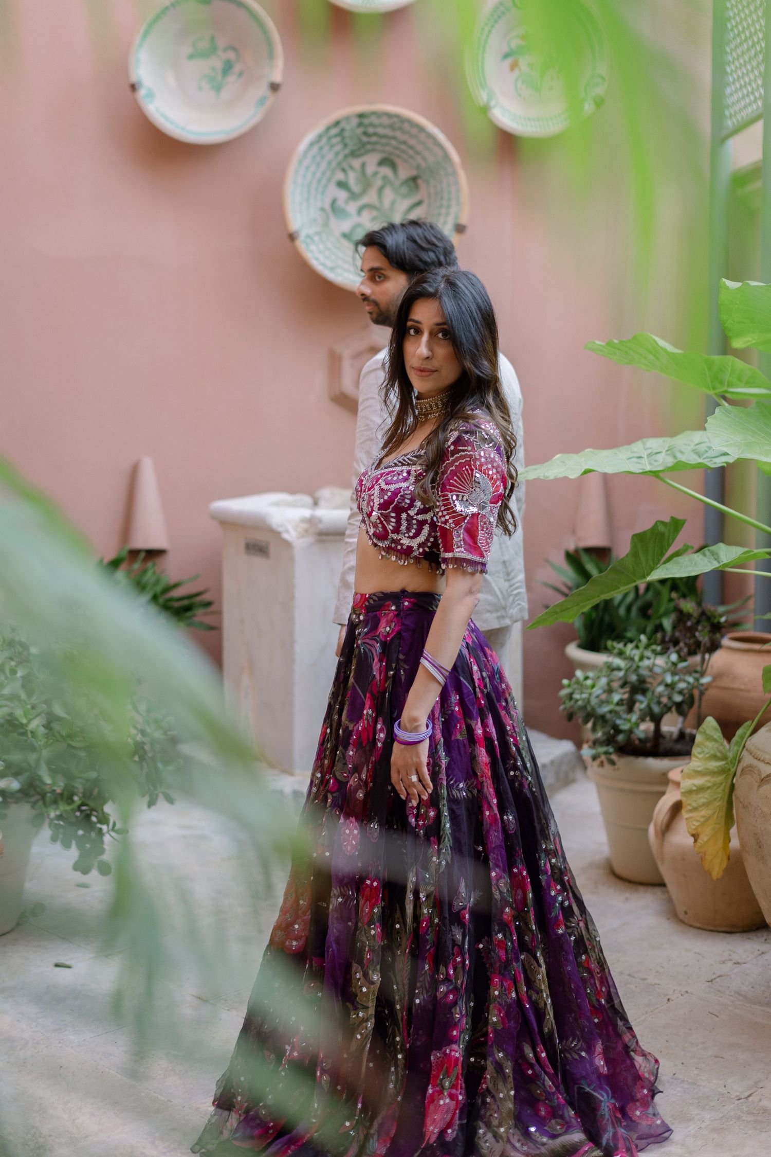 Elina looking towards the camera through leaves beside a pink wall with decorative plates.