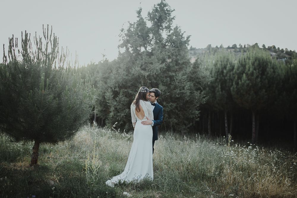 Two people share a romantic moment in their wedding attire on a misty hillside surrounded by tall evergreen trees.