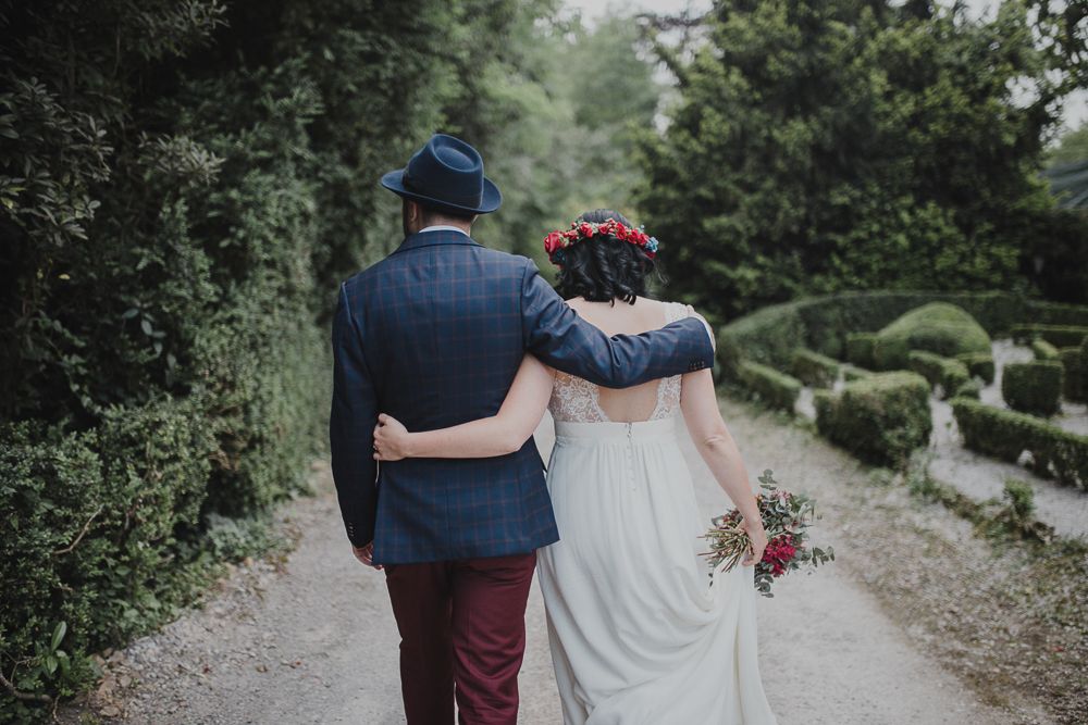 A couple walking down a garden path in wedding attire with greenery surrounding them.