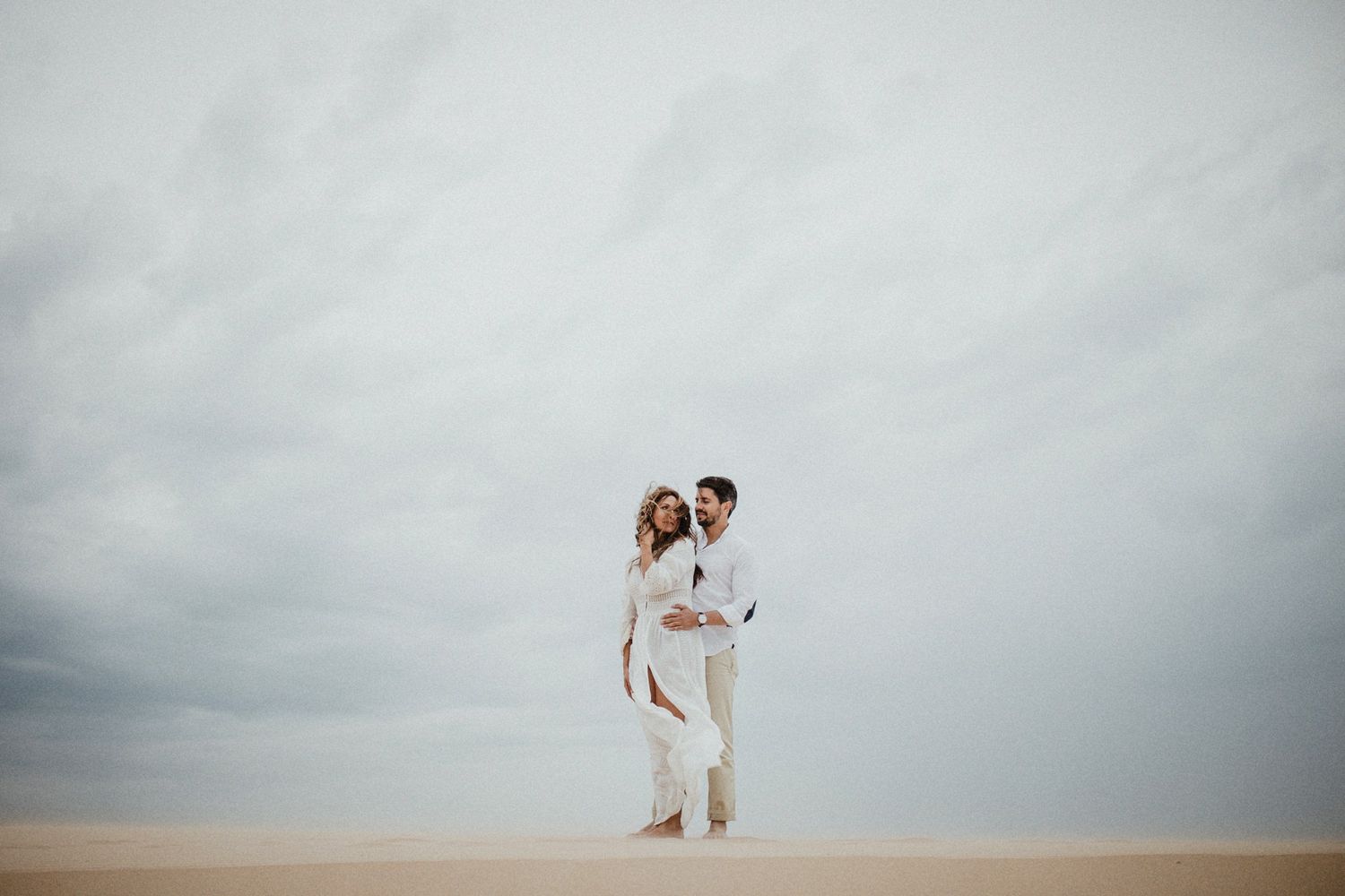 Couple in white outfits standing together on sandy dunes against moody grey sky.