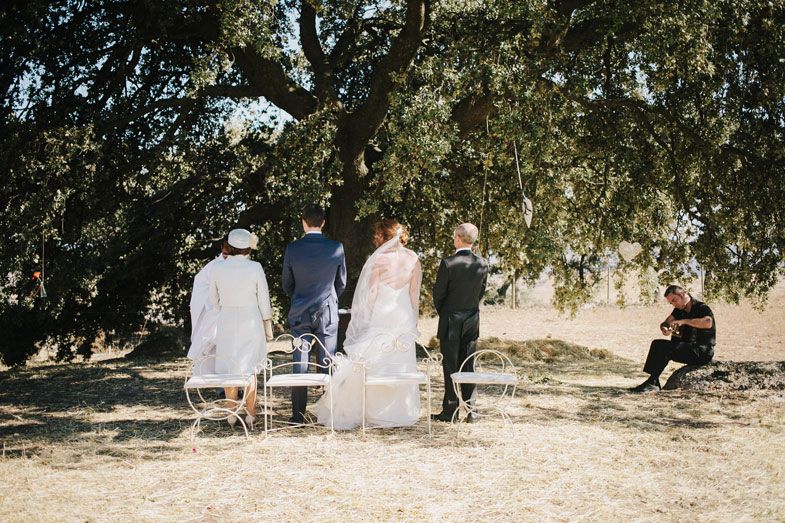 An outdoor wedding ceremony setup under a large tree with guests seated on chairs.