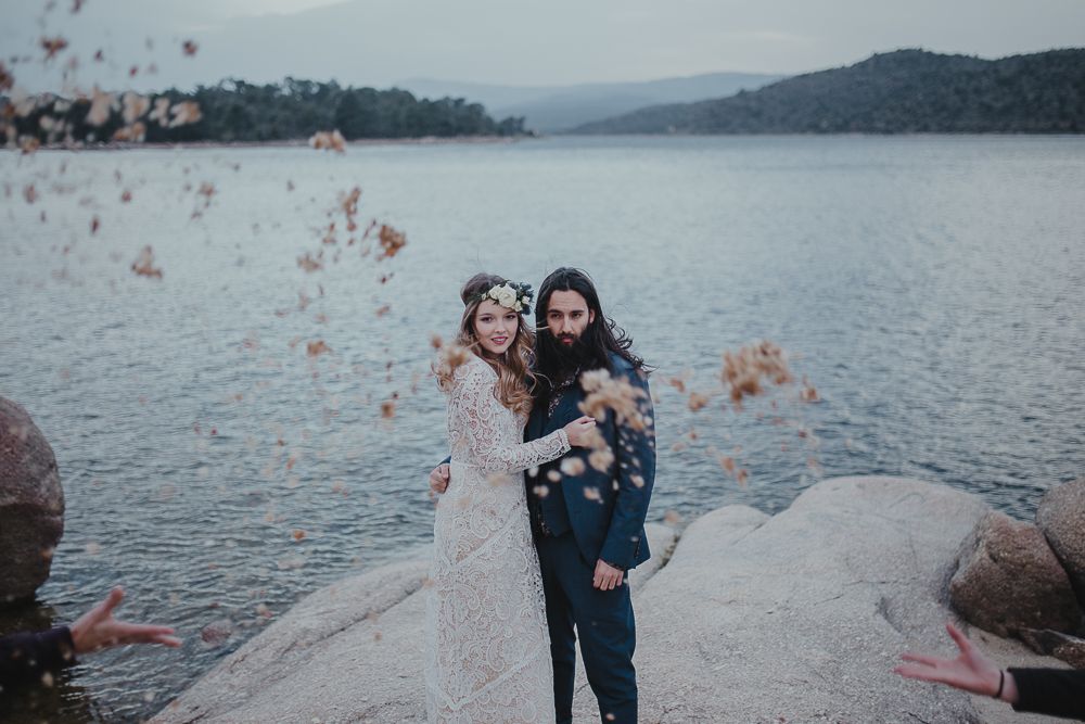 Couple celebrating with colorful smoke bombs by a serene lake surrounded by mountains.