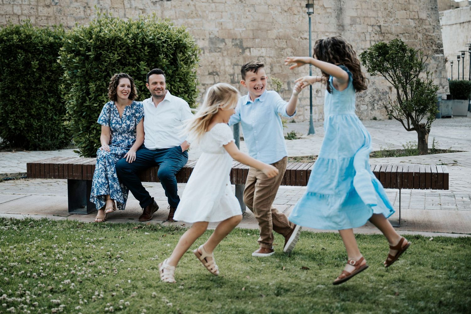 Family of four enjoying outdoor fun, parents watching children dance and play in a sunny park near a stone wall.