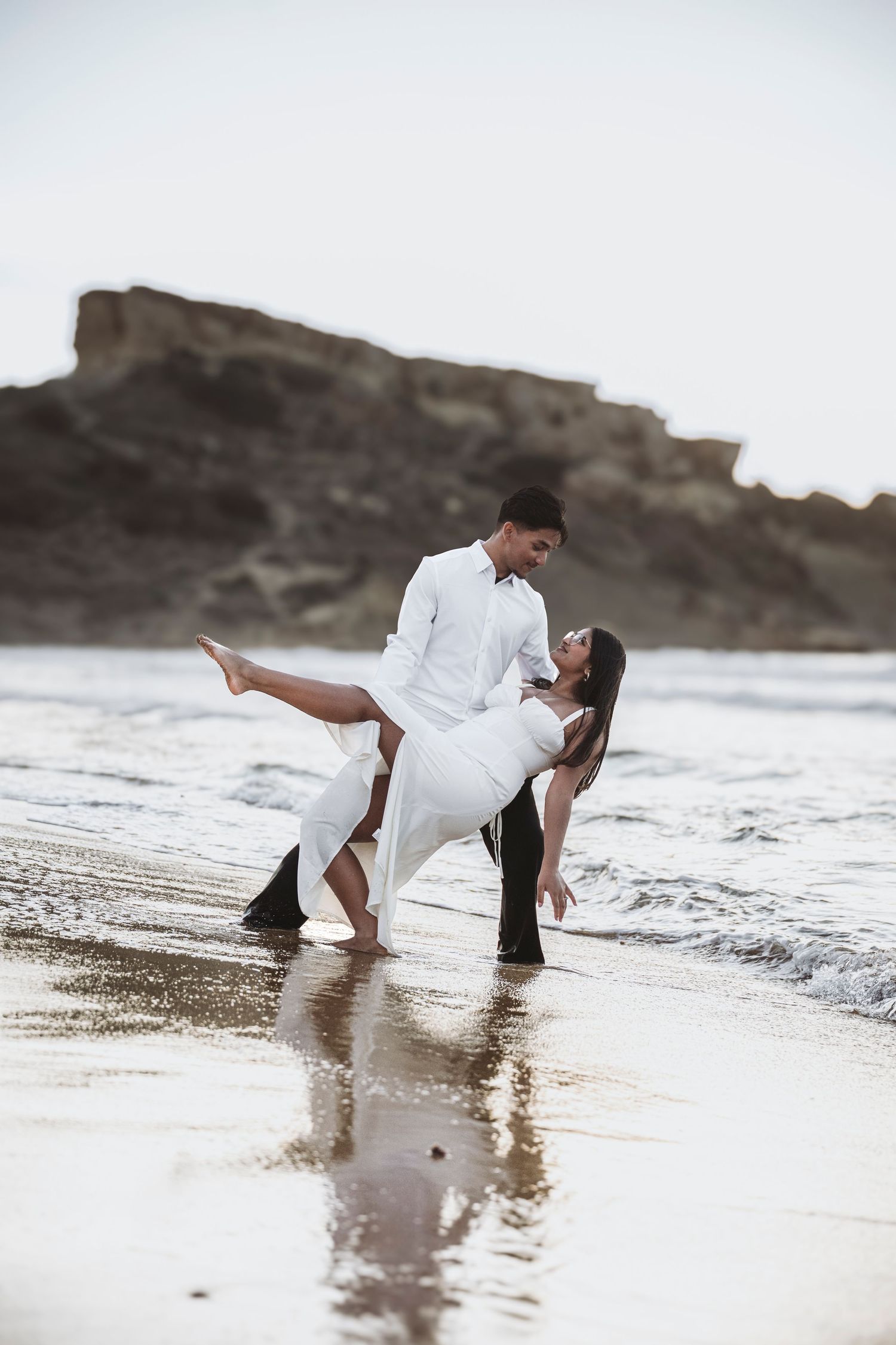 A romantic couple in white clothing plays in the ocean waves at the beach during a photo session.