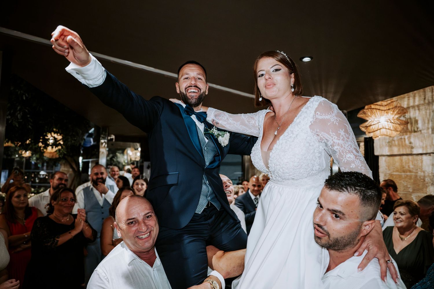 Bride and groom lifted on shoulders by guests during joyful wedding reception celebration indoors.