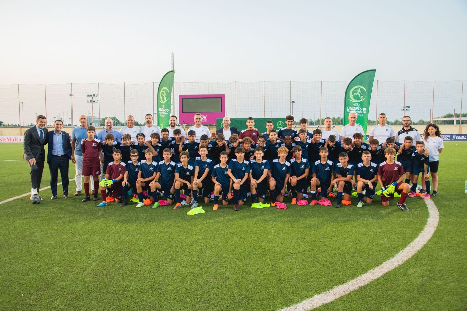 Large group of athletes in navy uniforms pose together on a soccer field with green banners in the background.