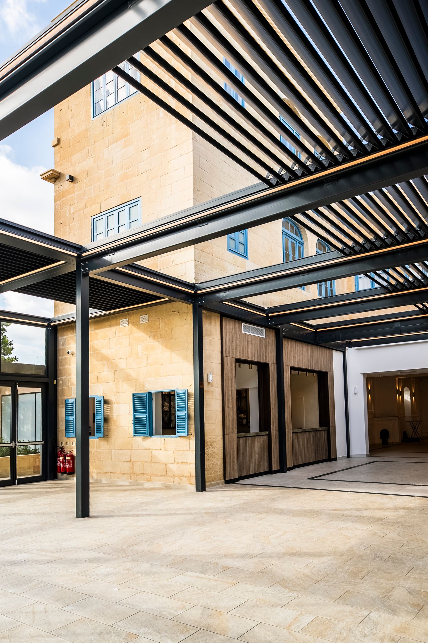 Modern covered walkway with black pergola beams casting shadows on stone tile flooring.