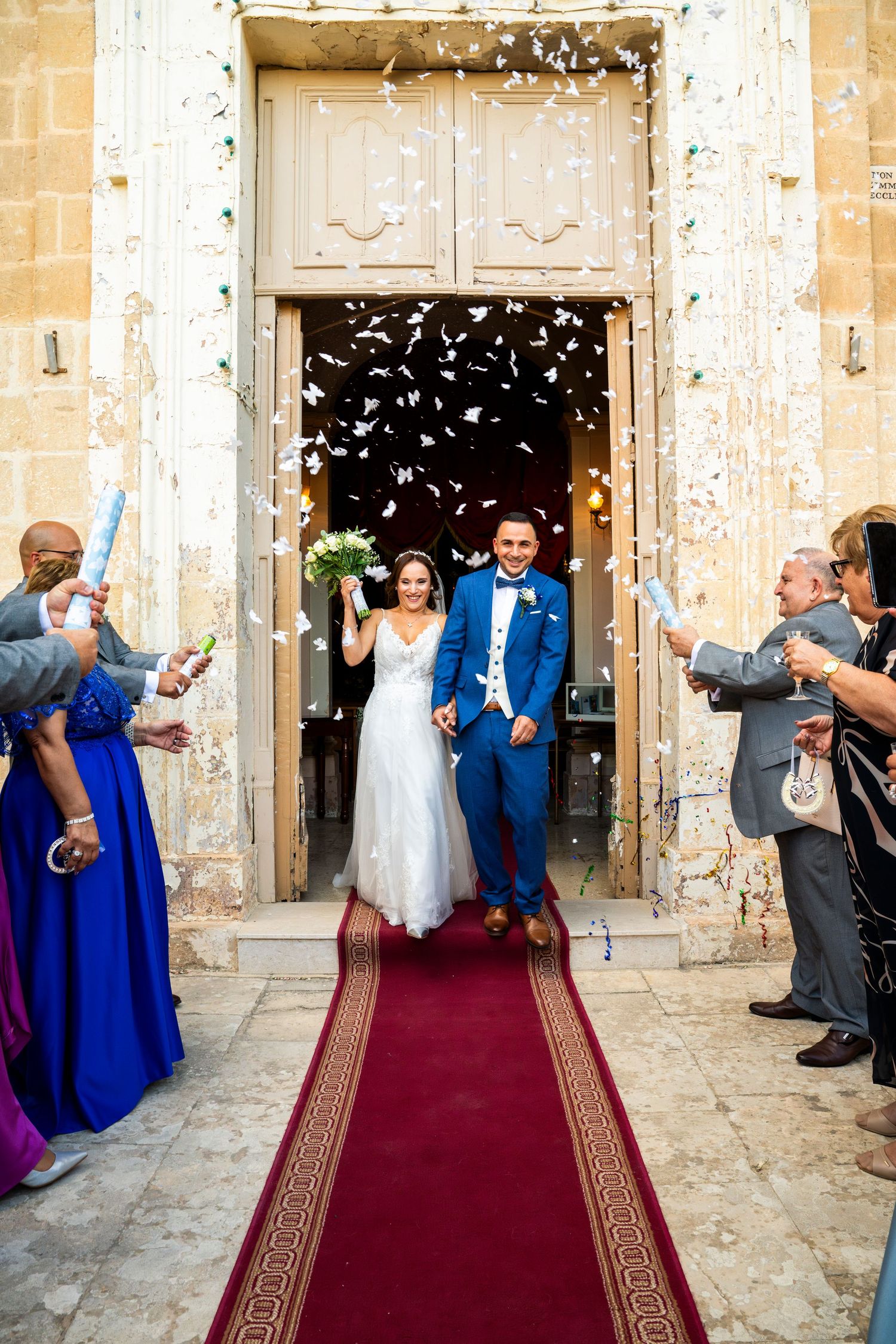 Newlywed couple exits church on red carpet as guests shower them with confetti during joyful wedding celebration.