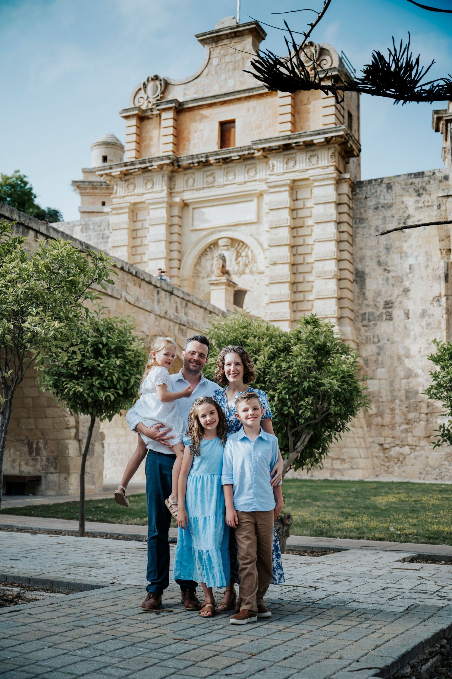 Family of five posing in front of historic stone gate in Malta, wearing coordinated blue and neutral outfits.