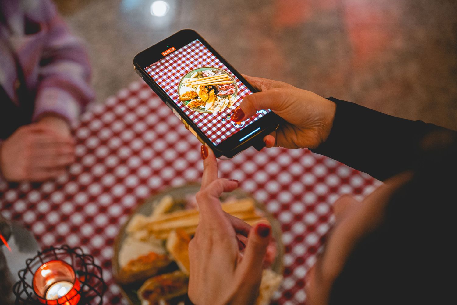 Hands holding smartphone taking photo of french fries on red checkered tablecloth.