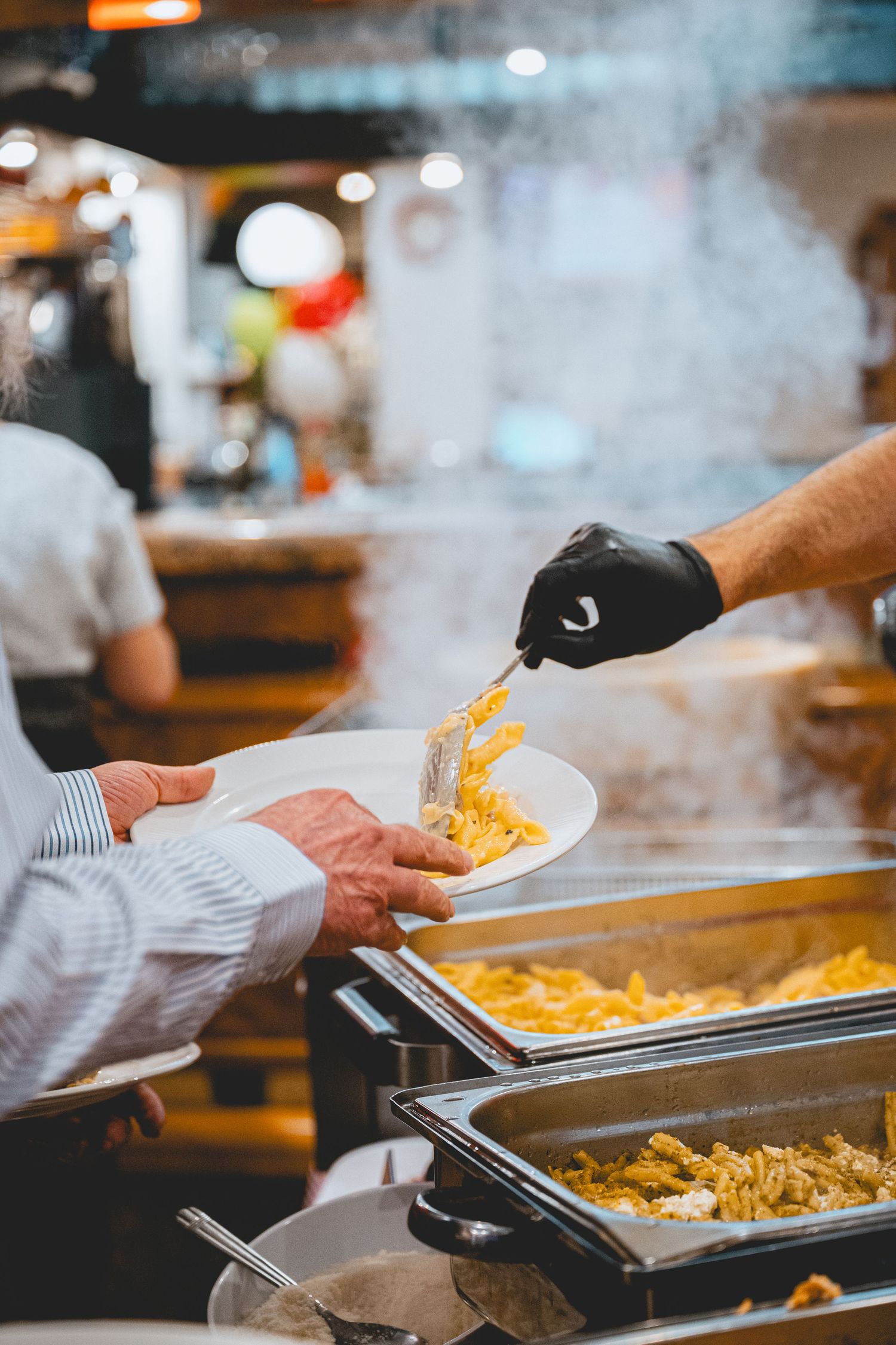 Chef serving hot food from steam trays in commercial kitchen wearing black gloves.