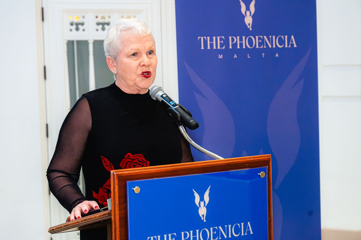 Speaker presents at The Phoenicia podium with blue branded backdrop in formal attire.