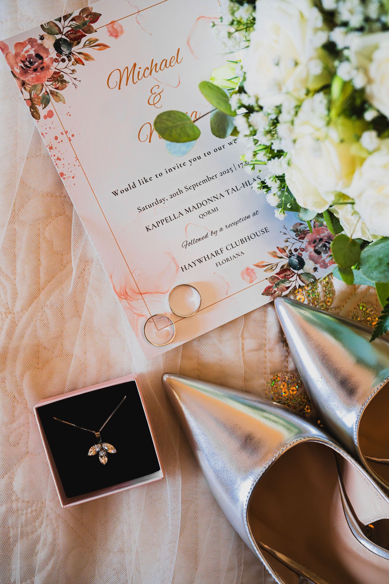 Wedding details including floral invitation, rings, and silver accessories on light fabric.