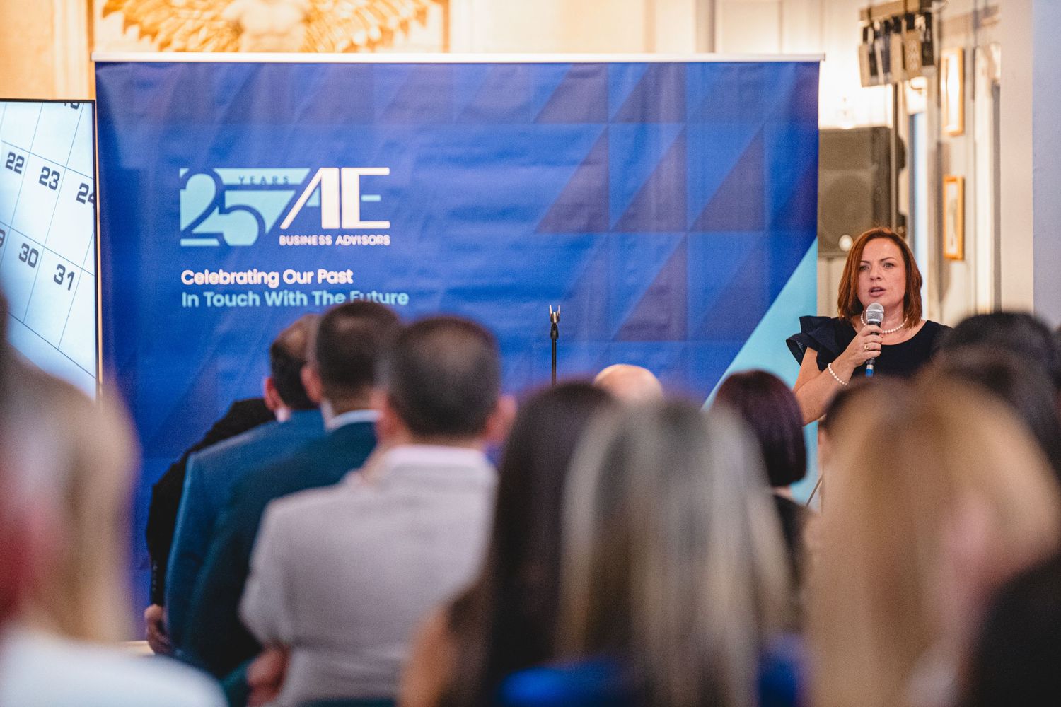 A business presentation taking place in an ornate room with a blue branded backdrop and attentive audience.
