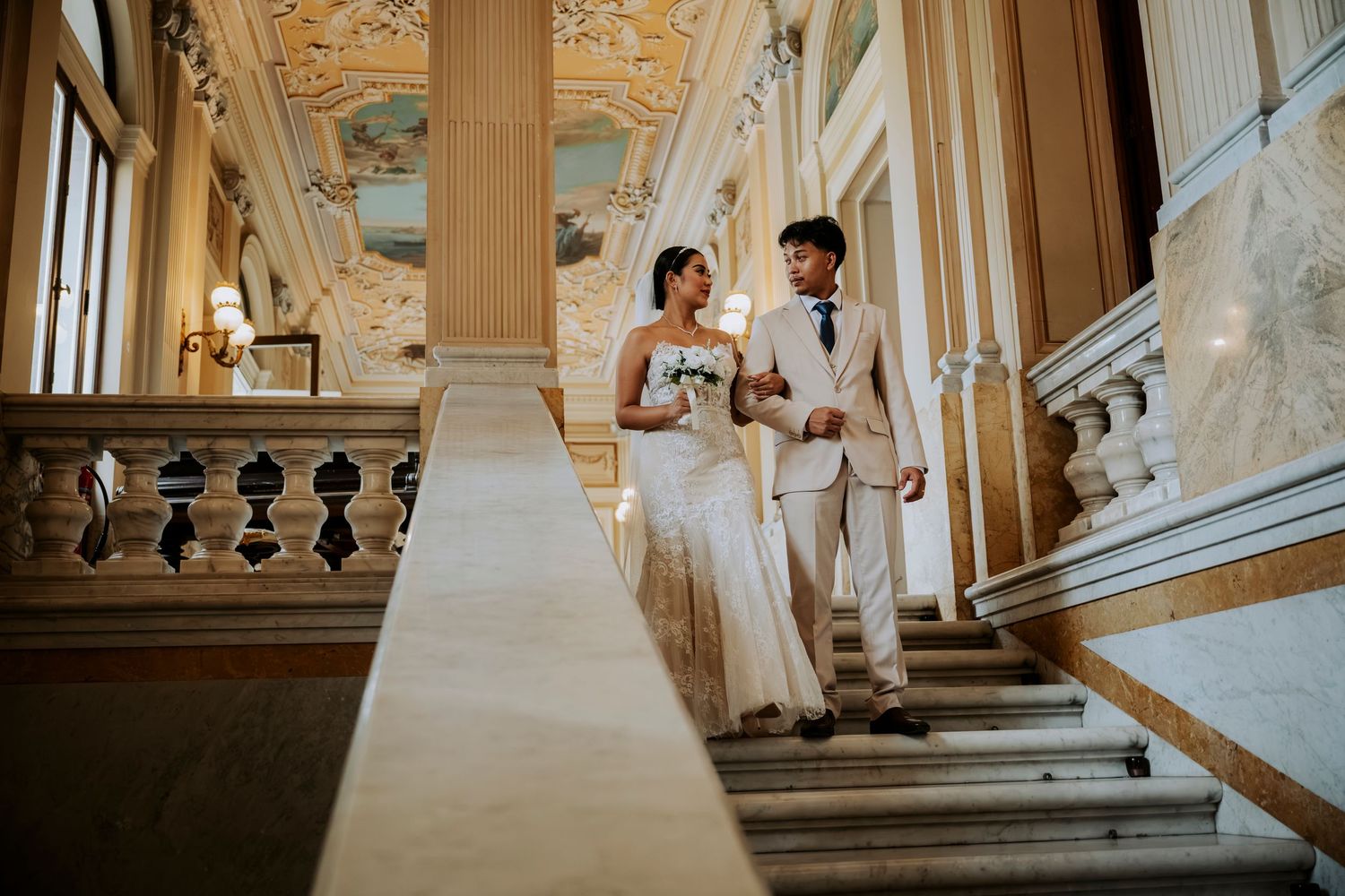 Elegant bride and groom walking down grand staircase in ornate wedding venue with golden ceiling.