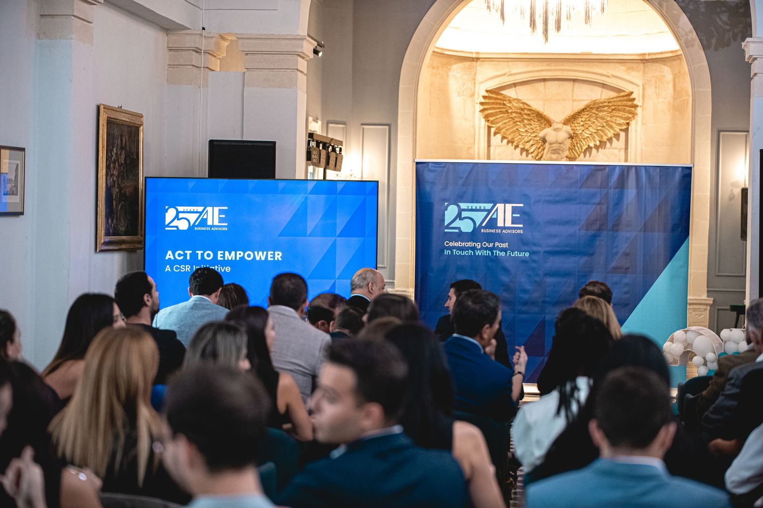 Conference attendees gather in an elegant room with blue banners and arched doorways for a professional event.