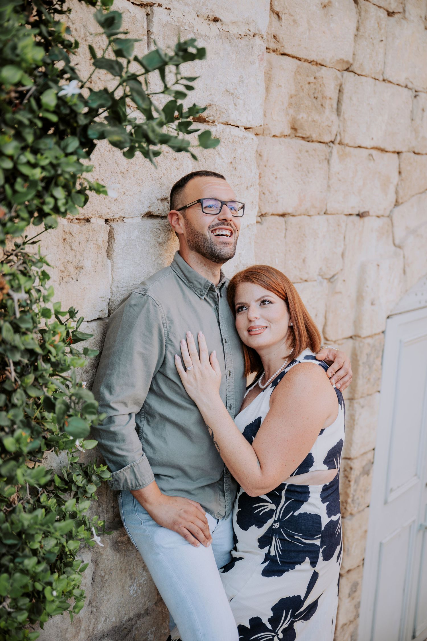 Happy couple posing against a stone wall outdoors, woman in floral dress embracing smiling man in glasses.