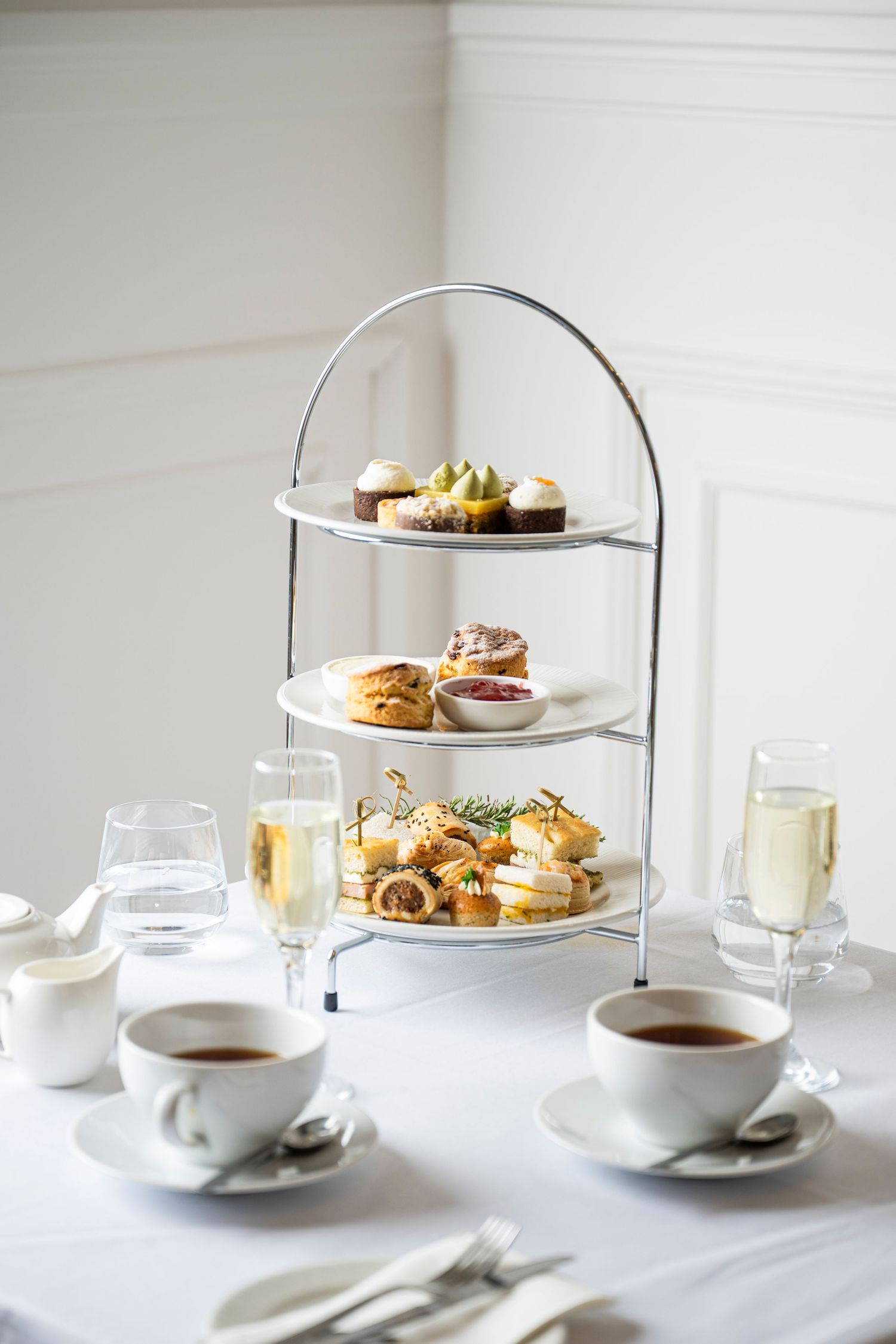 Elegant three-tier afternoon tea stand with pastries, scones, and sweets, served with champagne and tea cups on white linen.