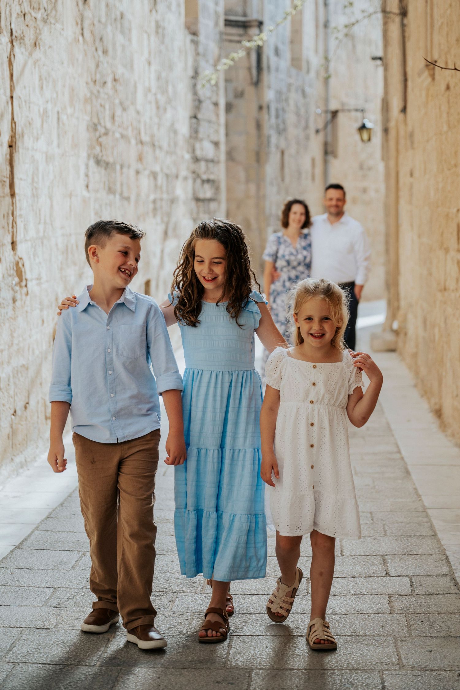 Three children walking and laughing through a narrow stone alley, parents following behind in a European old town.