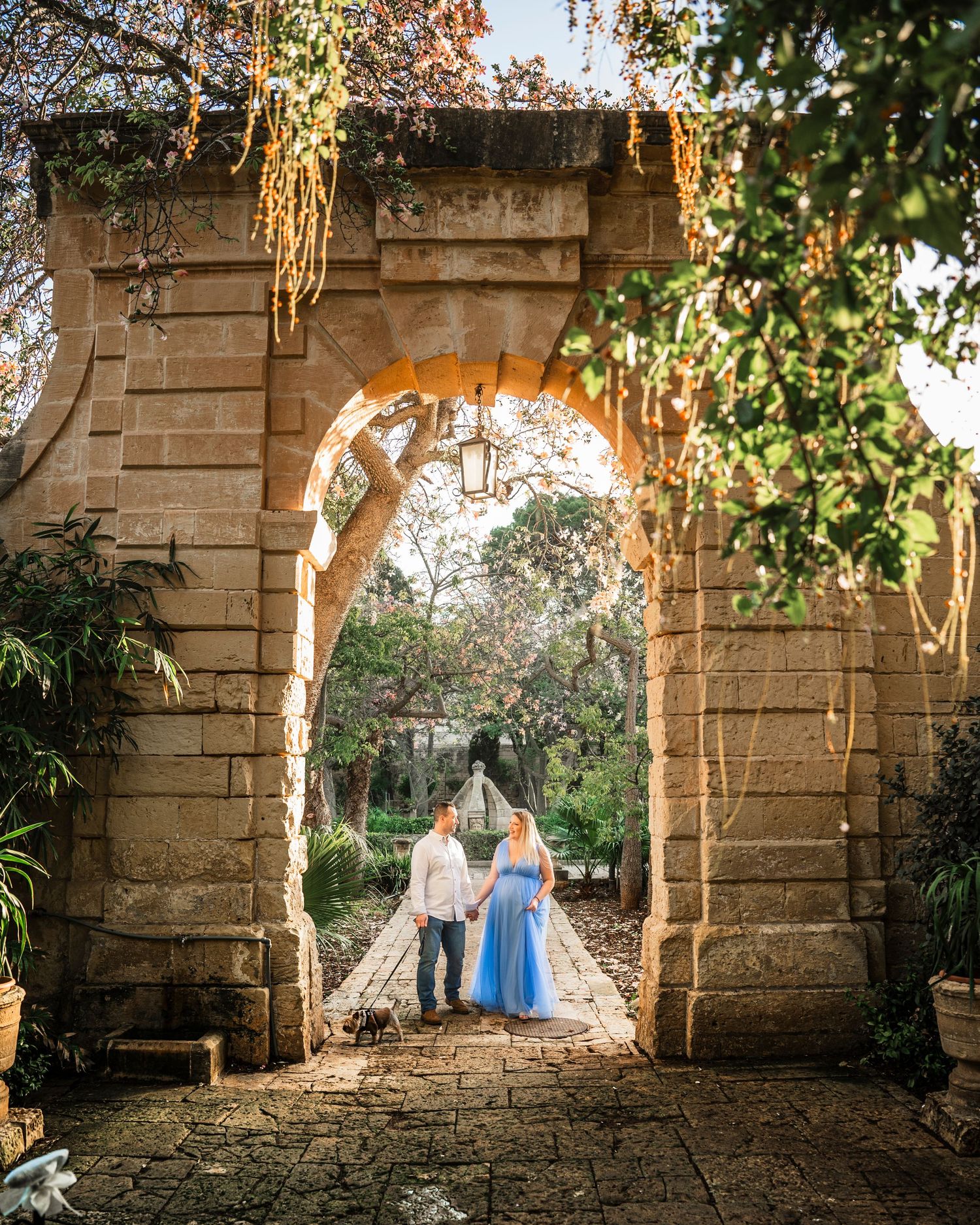 Couple holding hands under a stone arch in a lush garden, woman in blue dress, golden sunlight filtering through ivy.
