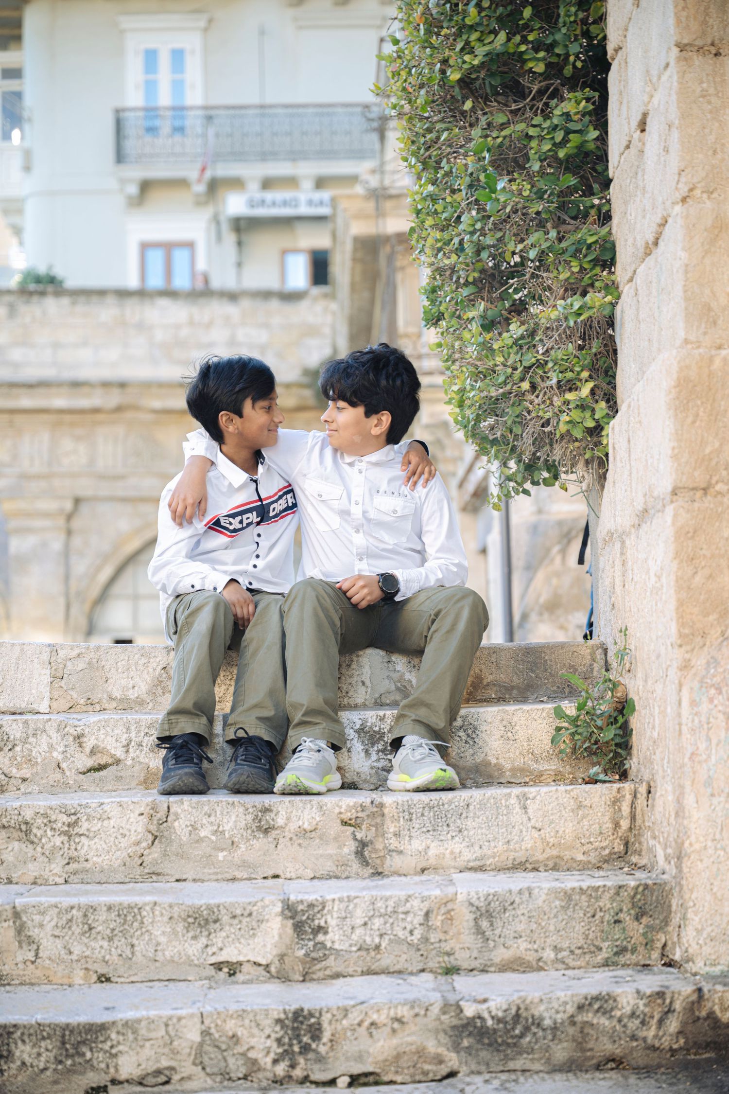 Two boys sitting on stone steps with arms around each other, smiling, with historic buildings in the background.