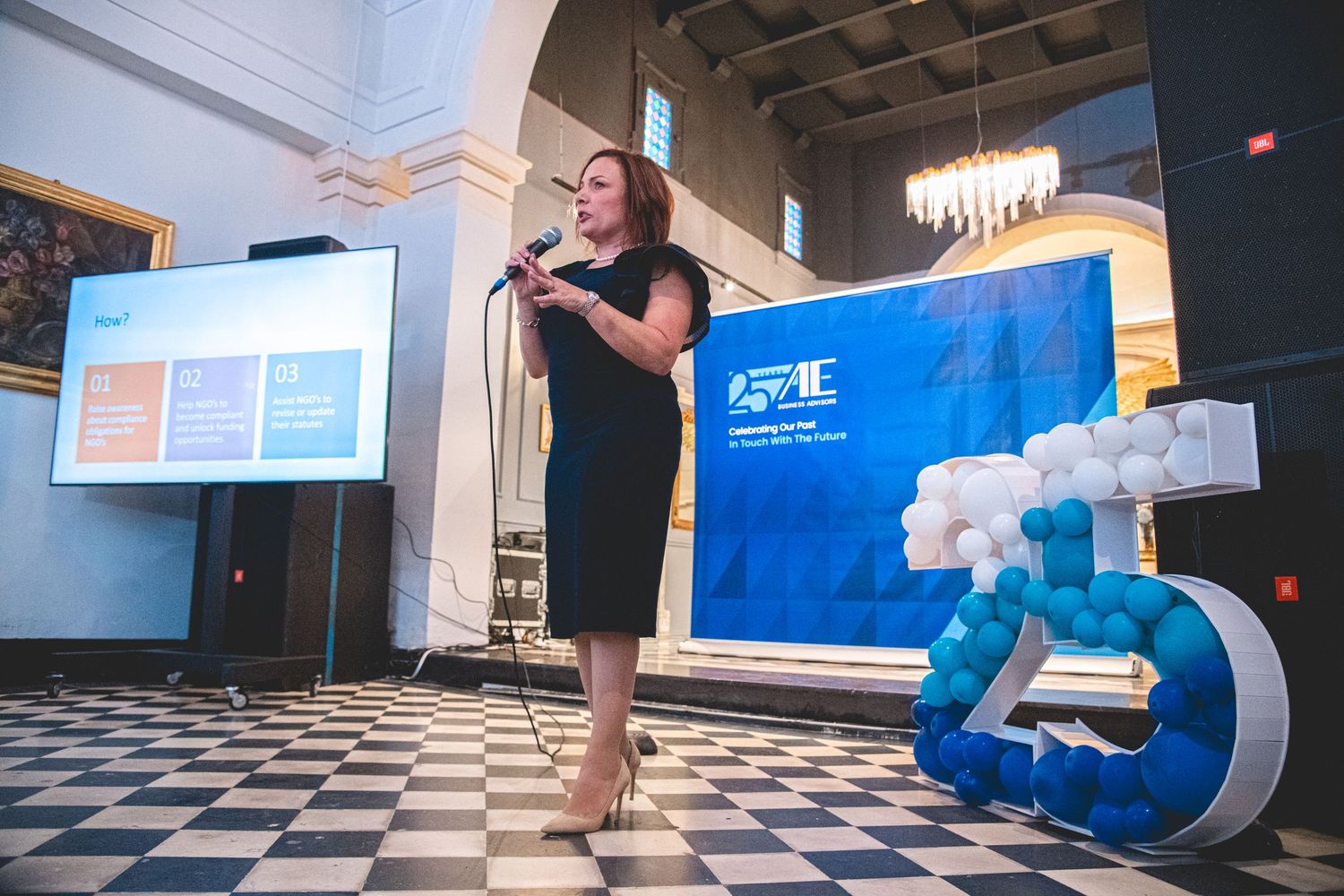 A presenter speaks at a podium with a blue branded backdrop and decorative 25 logo on a checkered floor.