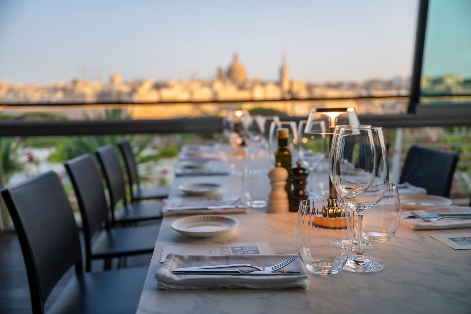 Elegant restaurant dining tables set with white linens and wine glasses against a scenic outdoor view at sunset.