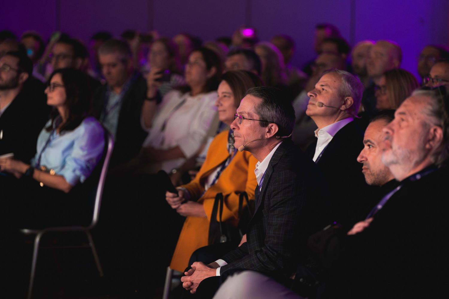 Business conference audience watching presentation in darkened auditorium with purple lighting.