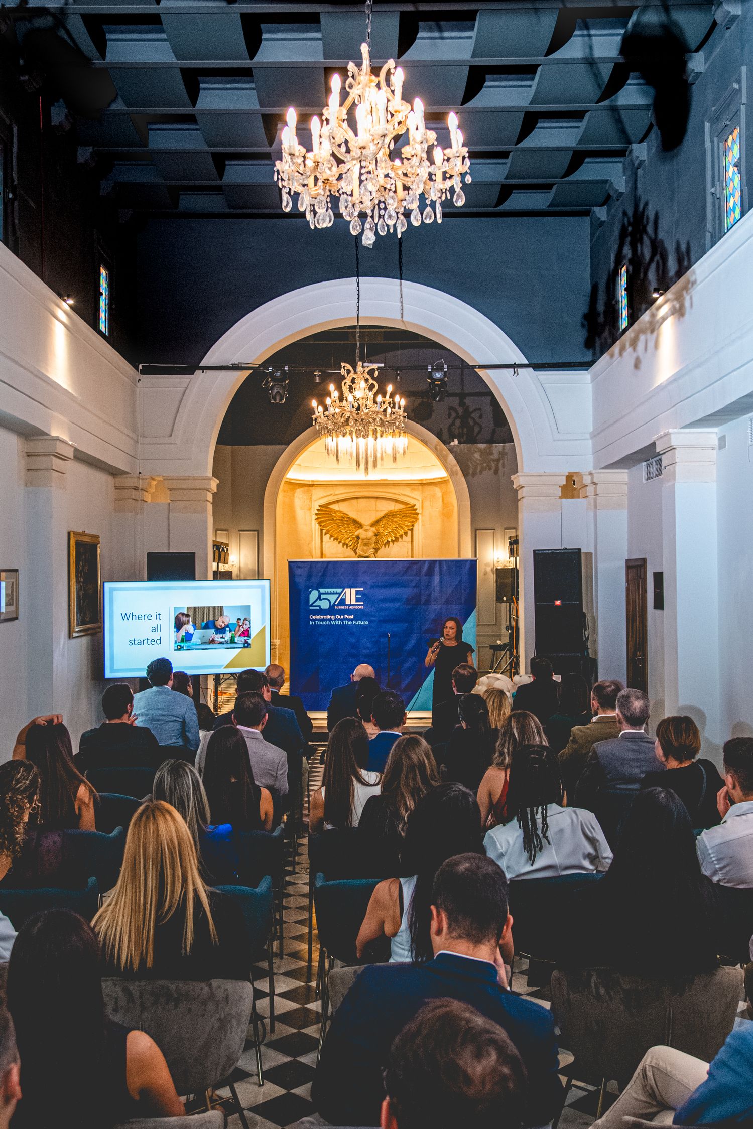 Conference or event space with chandeliers and audience seated in rows.
