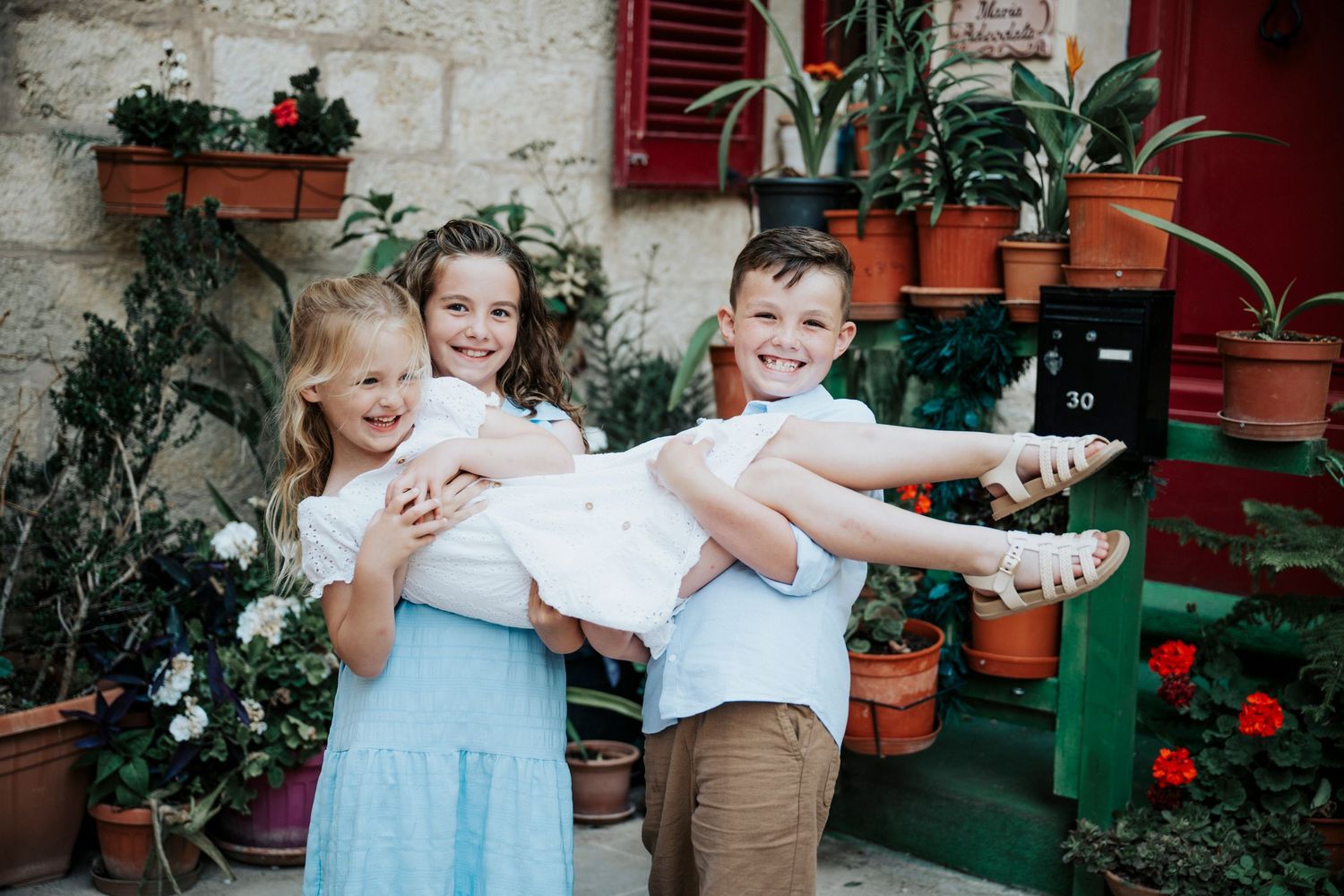 Three happy children pose playfully outdoors among colorful potted plants, two girls and a boy in white tops.
