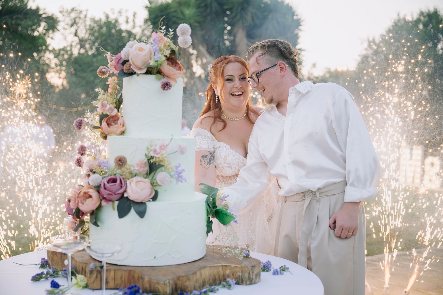 Bride and groom laughing while cutting a floral wedding cake surrounded by sparkling fireworks outdoors.