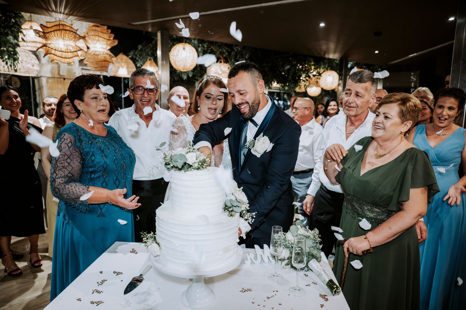 Two grooms cutting a white three-tier wedding cake surrounded by smiling guests in elegant attire at a reception.