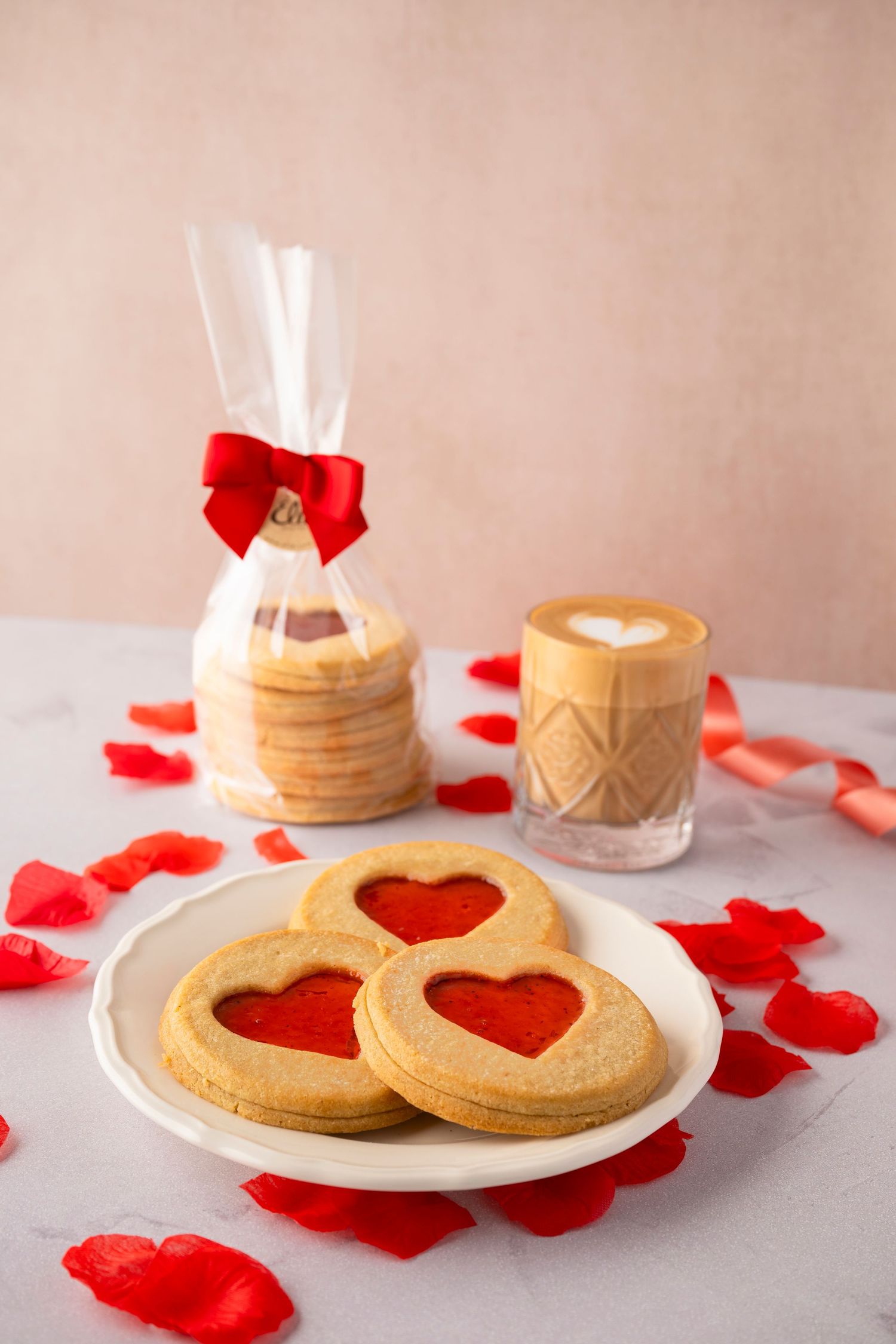 Heart-shaped Linzer cookies with red jam filling on white plate with coffee and rose petals for Valentine's Day celebration.