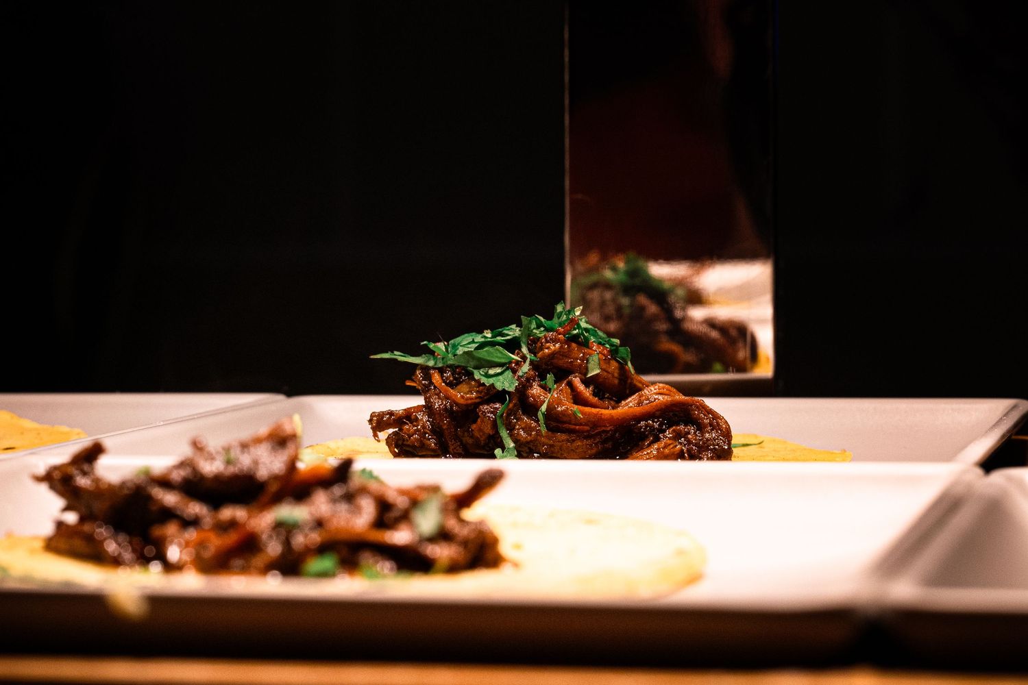 Gourmet meat dish plated with herbs on white square plate in dark lighting.