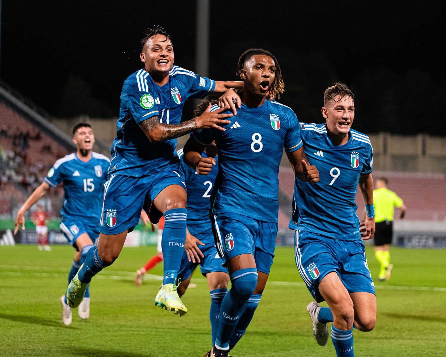 Players in blue jerseys embrace in celebration during nighttime soccer match.
