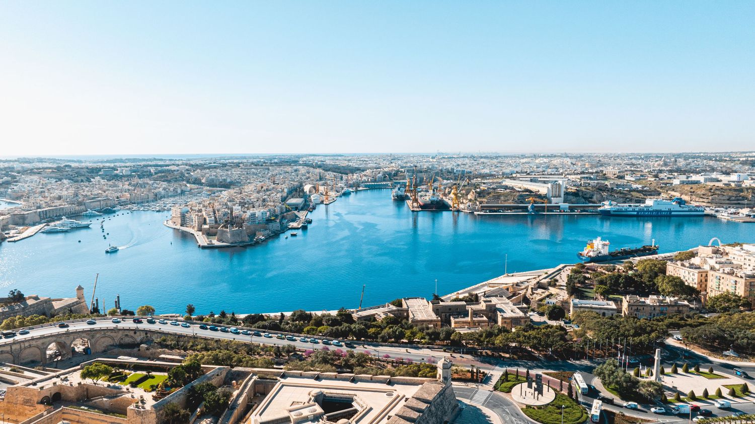 Aerial panorama of Valletta's harbor and historic cityscape against bright blue Mediterranean waters.