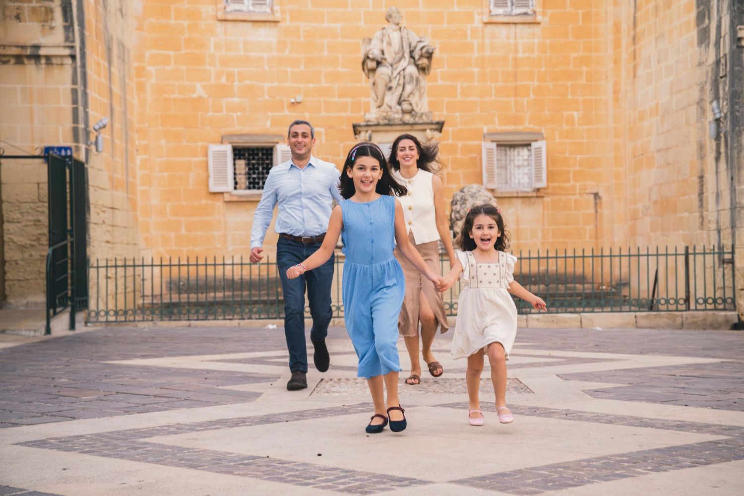 Family of four joyfully running in a sunlit European plaza with a stone statue and historic building backdrop.