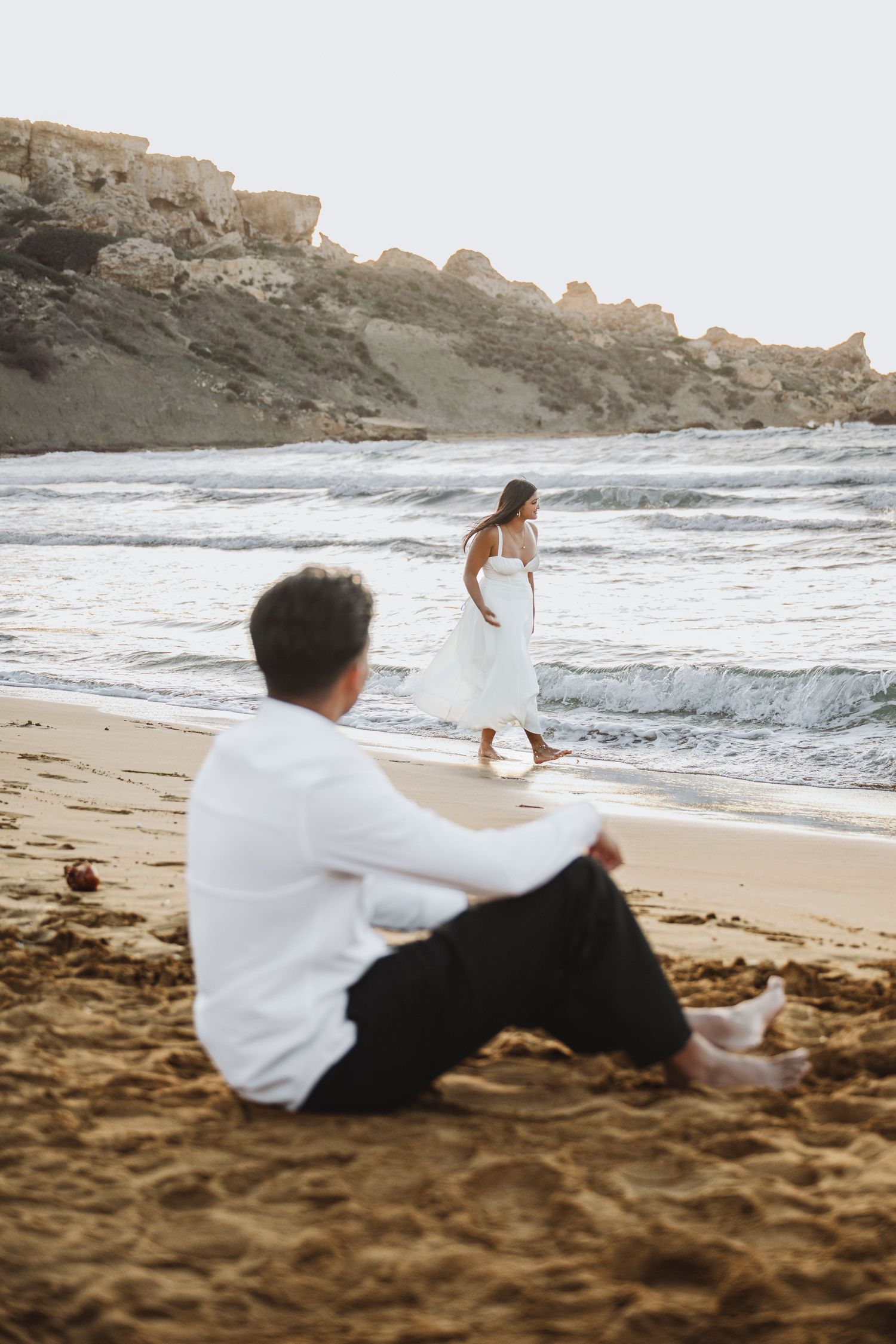 Couple playing and walking along shoreline with dramatic coastal cliffs at sunset.