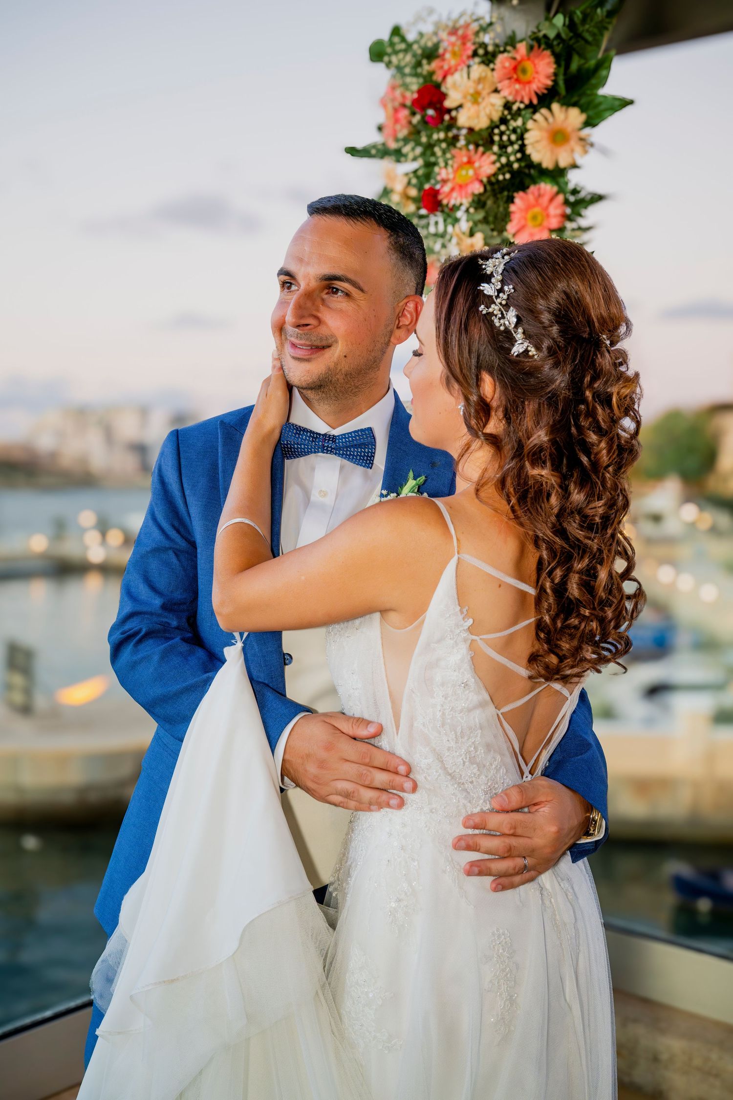 Newlyweds embrace by harbor with bride in white dress and groom in blue suit.