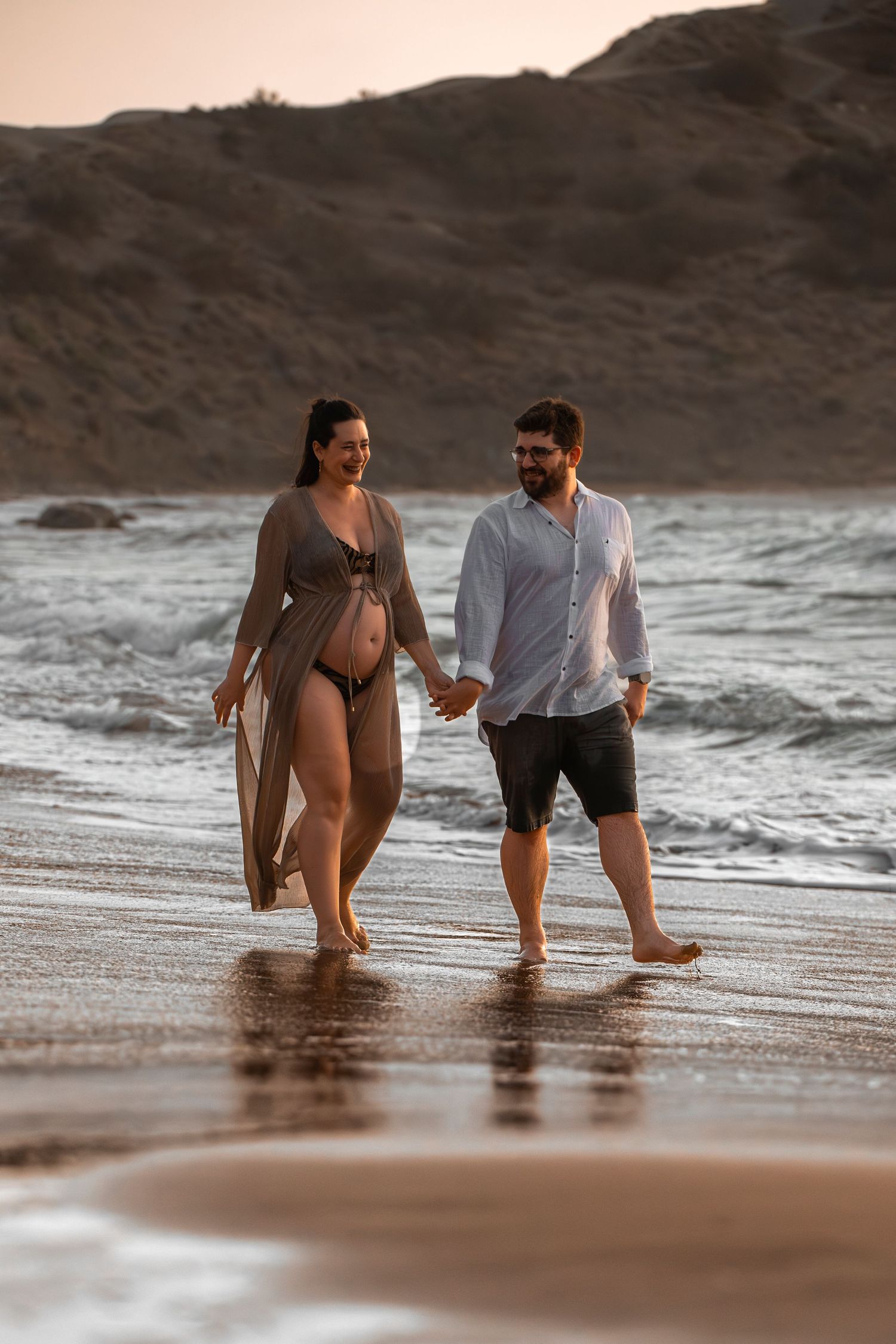 Pregnant couple holding hands walking barefoot on sandy beach at sunset with ocean waves and rocky cliffs in background.