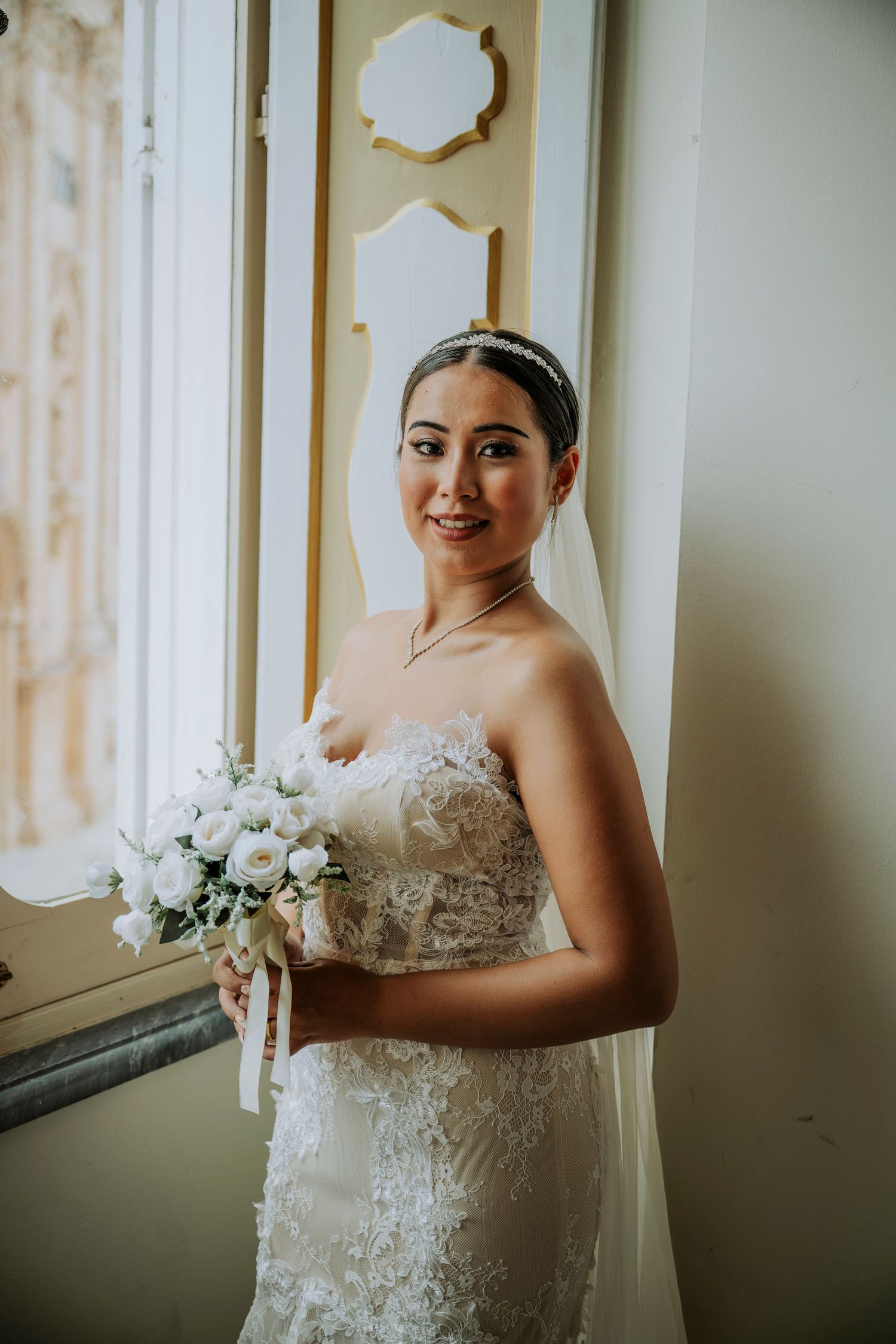 Bride in lace strapless gown holding white bouquet, wearing veil and tiara, standing by window.