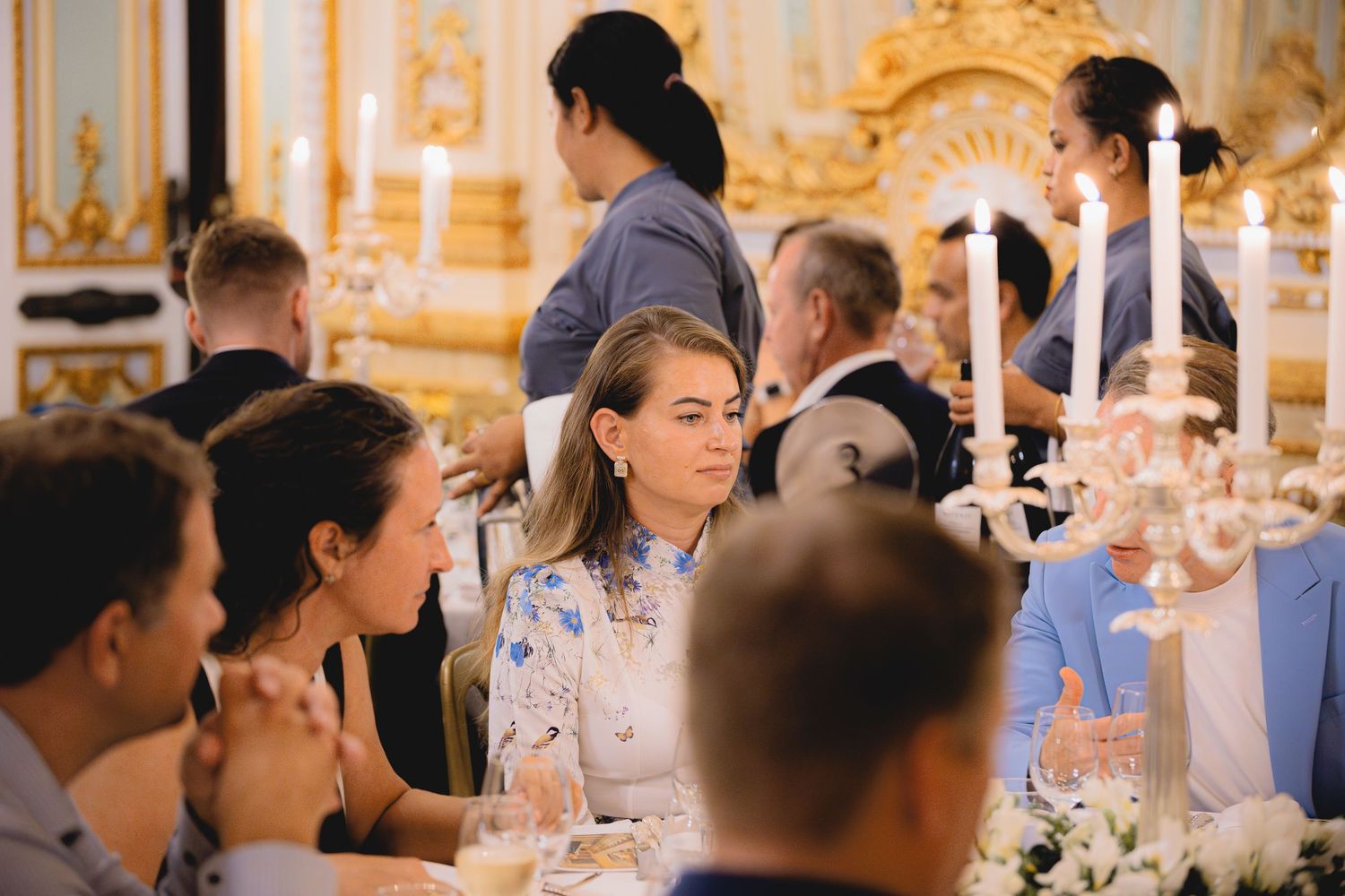 Formal dinner party with guests seated at long tables with white candles in an ornate gold room.