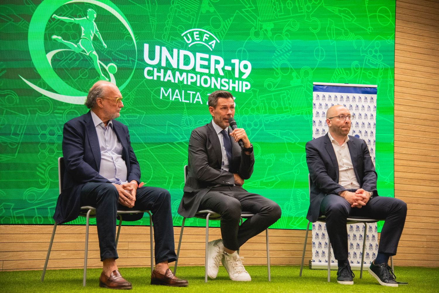 Three speakers seated on stage at UEFA Under-19 Championship event with green branded backdrop.