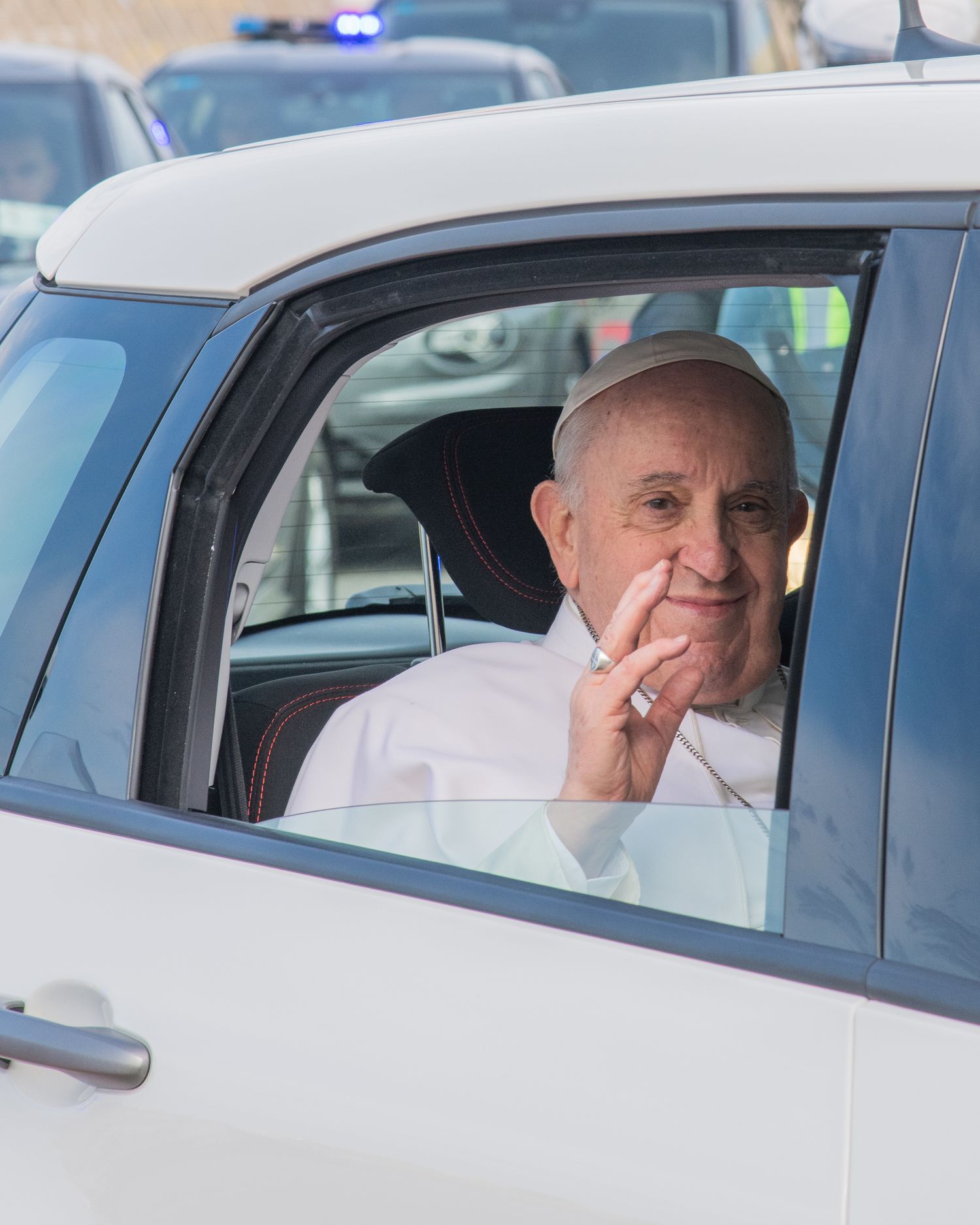 Religious figure waves from inside a white vehicle during official transport.