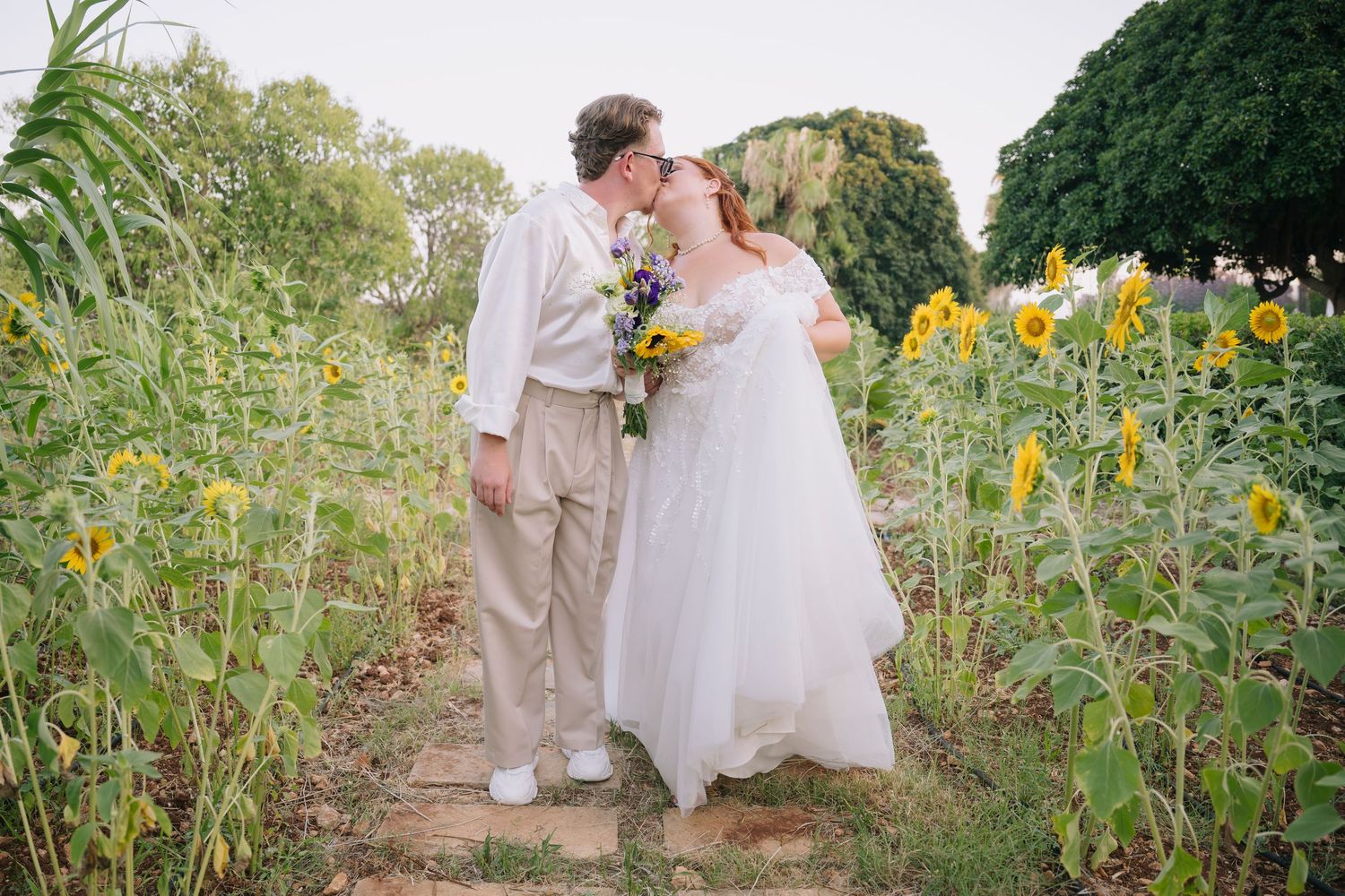 Bride and groom kissing in a sunflower field, bride in white gown holding yellow and blue bouquet.