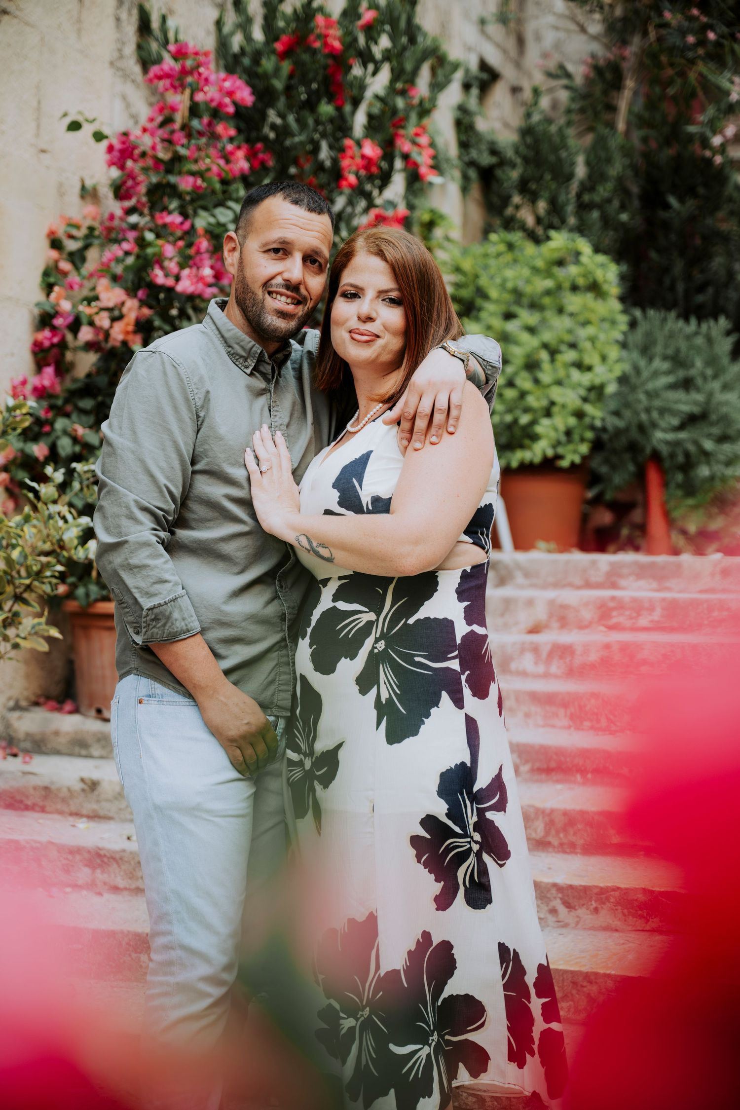 Couple embracing on pink steps surrounded by lush greenery and vibrant pink bougainvillea flowers.