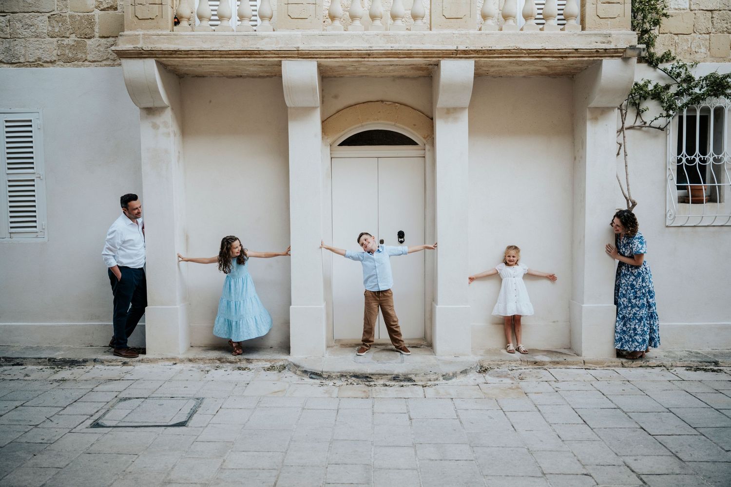Family of five posing with outstretched arms against a white Mediterranean building facade on a sunny day.