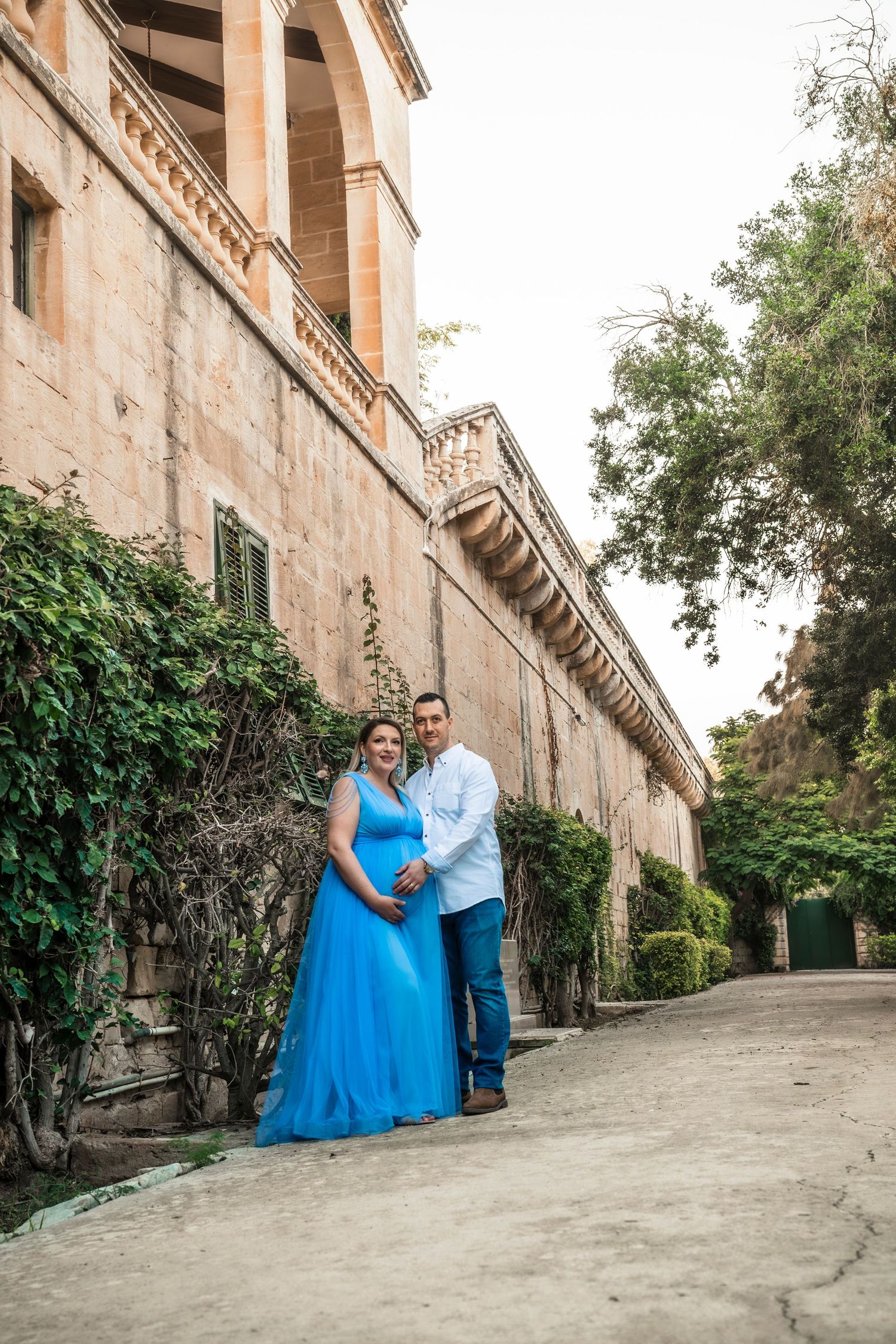Pregnant couple posing in front of a historic stone building with ivy, woman in blue maternity gown, man in white shirt.